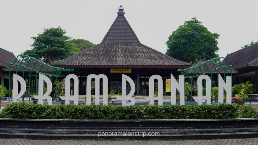 Large white letters spelling "PRAMBANAN" in front of a traditional Javanese joglo-style building at the park entrance.