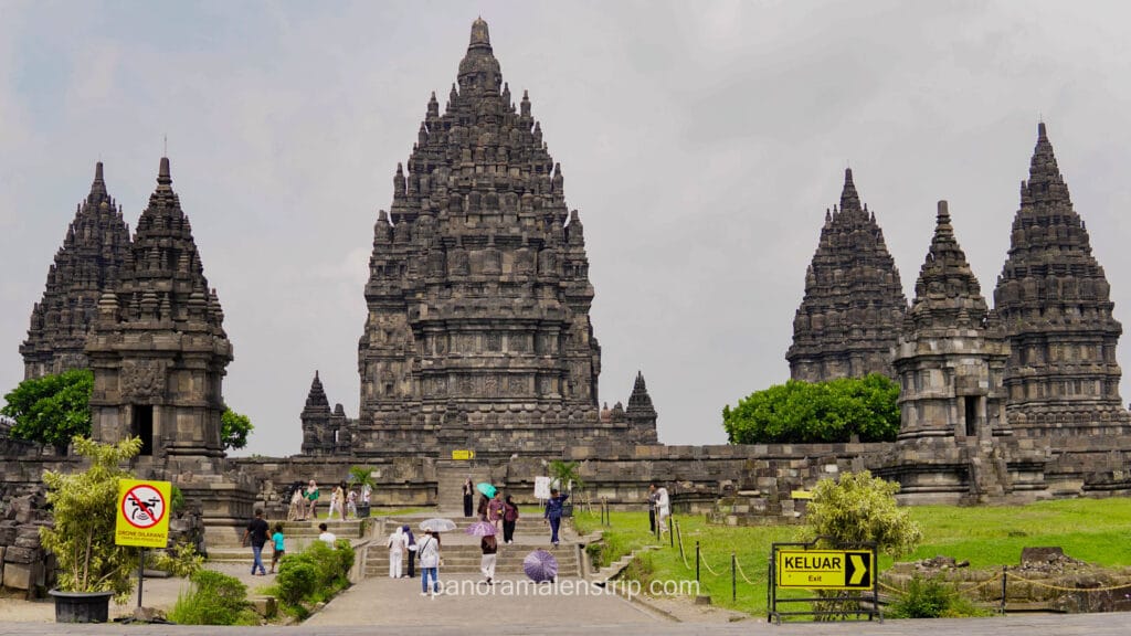 Wide shot of the Prambanan Temple complex with tourists walking on paved paths towards the three main shrines.