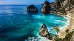Panoramic view of Diamond Beach in Nusa Penida, Bali, featuring dramatic limestone cliffs, pointed rock formations in clear turquoise water, and a pristine white sand beach fringed with palm trees.