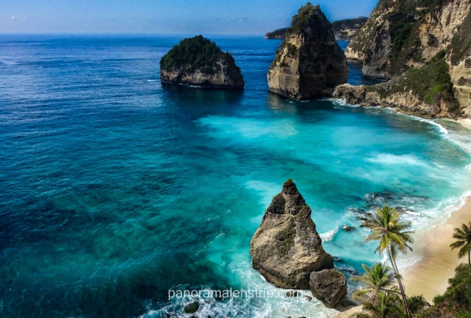 Panoramic view of Diamond Beach in Nusa Penida, Bali, featuring dramatic limestone cliffs, pointed rock formations in clear turquoise water, and a pristine white sand beach fringed with palm trees.