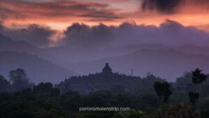 A silhouetted view of the ancient Borobudur temple complex in Central Java, Indonesia, nestled within a dramatic, layered landscape of misty purple and blue mountains. A multi-colored sunrise sky with pink, orange, and purple clouds and low-hanging mist is visible above, with a dense forest of tropical trees in the foreground and a small communication tower to the right of the main stupa.