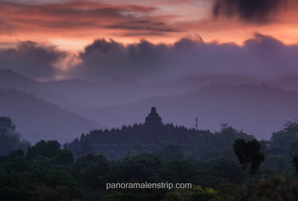 A silhouetted view of the ancient Borobudur temple complex in Central Java, Indonesia, nestled within a dramatic, layered landscape of misty purple and blue mountains. A multi-colored sunrise sky with pink, orange, and purple clouds and low-hanging mist is visible above, with a dense forest of tropical trees in the foreground and a small communication tower to the right of the main stupa.