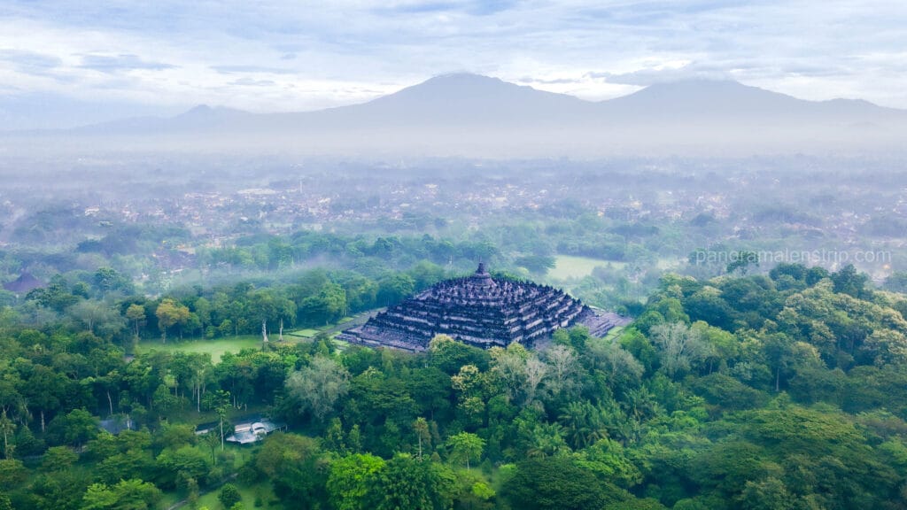 An aerial photograph of the massive Borobudur temple, a complex stepped pyramid of stone stupas, nestled in a dense green Indonesian jungle. A misty valley spreads beyond, leading to two large, cloud-shrouded mountains under a soft, overcast morning sky.