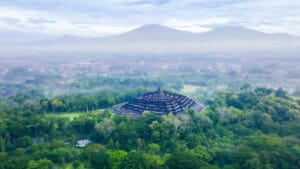 An aerial photograph of the massive Borobudur temple, a complex stepped pyramid of stone stupas, nestled in a dense green Indonesian jungle. A misty valley spreads beyond, leading to two large, cloud-shrouded mountains under a soft, overcast morning sky.