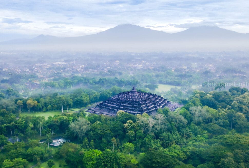 An aerial photograph of the massive Borobudur temple, a complex stepped pyramid of stone stupas, nestled in a dense green Indonesian jungle. A misty valley spreads beyond, leading to two large, cloud-shrouded mountains under a soft, overcast morning sky.