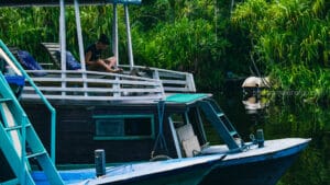 A woman sitting cross-legged and relaxing on the open upper deck of a traditional wooden klotok boat, floating on a jungle river with dense green vegetation in the background.