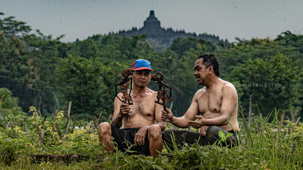 Two Javanese men, shirtless, sit in a field with a pair of leather Wayang Kulit shadow puppets, with the ancient stupas of Borobudur temple visible in the far background under a cloudy sky.