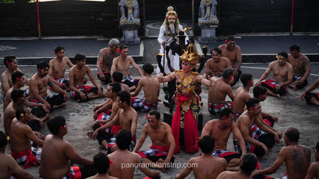 A dynamic performance of the traditional Balinese Kecak dance. A female dancer in an ornate, golden costume dances in the center of a large circle of seated, bare-chested men wearing checkered sarongs. A mythical masked figure in a white costume stands in the background near stone statues.