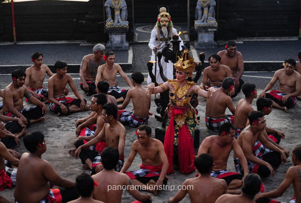 A dynamic performance of the traditional Balinese Kecak dance. A female dancer in an ornate, golden costume dances in the center of a large circle of seated, bare-chested men wearing checkered sarongs. A mythical masked figure in a white costume stands in the background near stone statues.