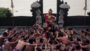 An authentic Balinese Kecak fire dance performance at the Uluwatu Temple cliffside. A central female dancer in an ornate gold headdress and traditional crimson and gold costume is hoisted by a male performer, surrounded by a large, bare-chested male chanting choir with raised hands, all in front of the iconic 'Candi Bentar' split gate and the ocean at sunset. The choir members wear traditional red and white checkered poleng cloth.