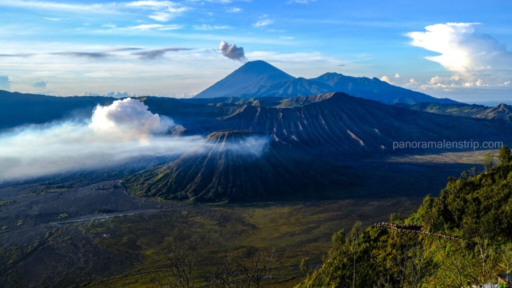 A high-angle panoramic view showing the Bromo volcanic complex, the Sea of Sand, and the towering peak of Mount Semeru erupting in the far distance under a bright blue sky.