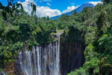 Scenic view of Tumpak Sewu Waterfall with the peak of Mount Semeru volcano in the background under a blue sky.