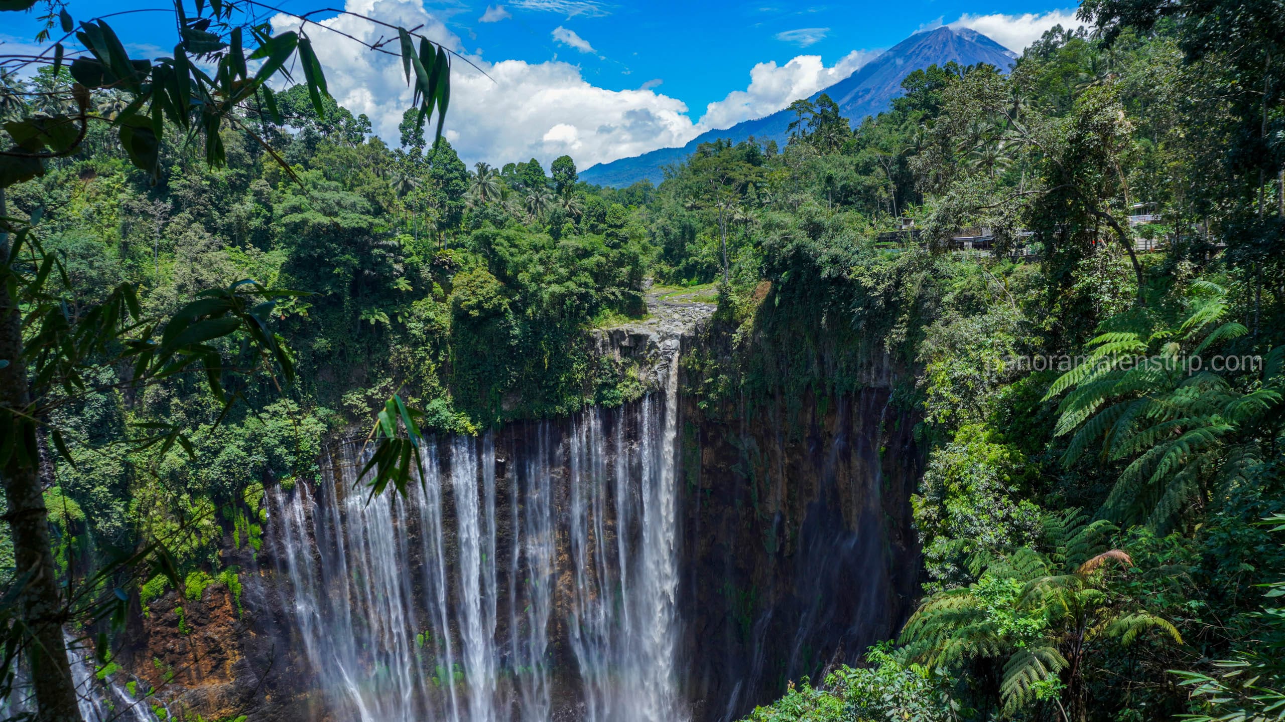 Scenic view of Tumpak Sewu Waterfall with the peak of Mount Semeru volcano in the background under a blue sky.