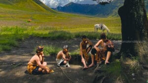 Four young Tenggerese boys wearing traditional batik sarongs and headbands (Udeng) sitting under a tree in the Bromo savanna; one boy is playing a violin while a white horse grazes in the background.
