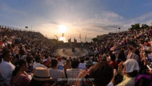 A large, diverse crowd of international tourists seated in a circular stone amphitheater at Uluwatu Temple in Bali, Indonesia, looking toward a central stage and a traditional Balinese split gate with a golden sunset over the ocean in the background.
