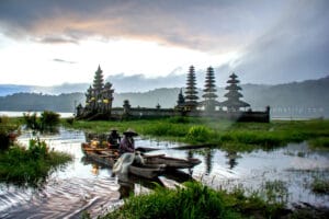 Traditional wooden boat with fishermen on Tamblingan Lake at sunrise with ancient temple and misty mountains, one of the most scenic bali photography spots.