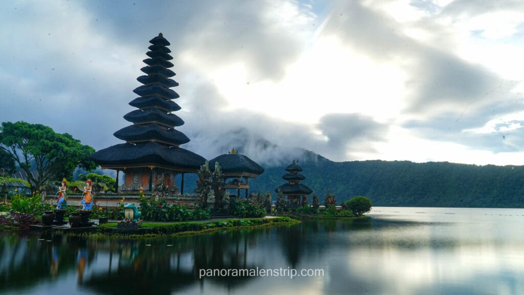 A beautiful view of the Pura Ulun Danu Beratan water temple in Bali, framed by bright red Canna lilies in the foreground. The temple's iconic multi-tiered thatched pagoda sits on a small island surrounded by calm lake water and colorful gardens under a cloudy sky.
