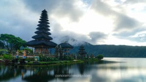 A beautiful view of the Pura Ulun Danu Beratan water temple in Bali, framed by bright red Canna lilies in the foreground. The temple's iconic multi-tiered thatched pagoda sits on a small island surrounded by calm lake water and colorful gardens under a cloudy sky.