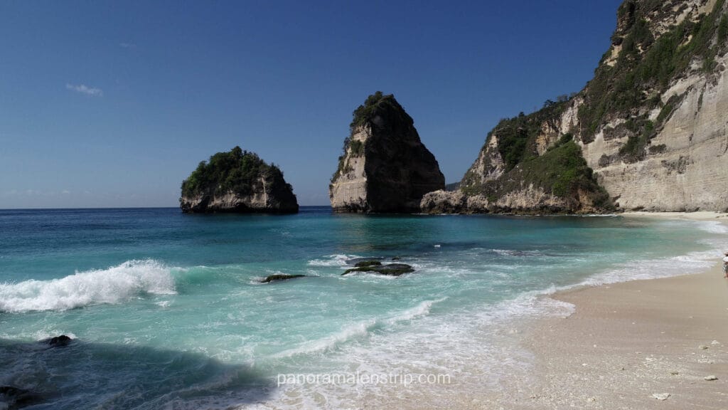 A breathtaking view of a pristine white sand beach in Nusa Penida with vibrant turquoise ocean water and white crashing waves. Two large, sheer limestone rock formations rise sharply from the sea, flanked by a massive, vegetation-covered cliff on the right under a clear blue sky.