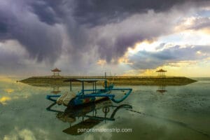 A blue traditional Balinese outrigger boat anchored in shallow, calm water at Sanur Beach. The mirror-like water reflects the boat and a dramatic sky, where dark storm clouds are pierced by striking golden sun rays shining down on two distant gazebos on a breakwater.