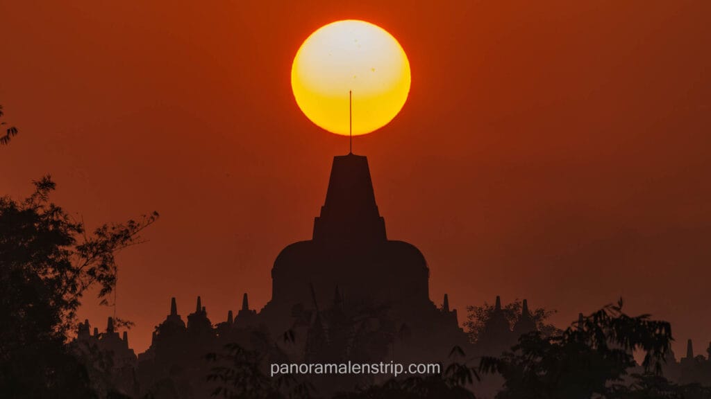 A stunning silhouette of the magnificent Borobudur temple complex in Java, Indonesia, against a fiery red-orange sunset sky. The large, golden sun orb is perfectly positioned directly above the central stupa peak, creating a dramatic halo effect. Dense tree branches are in the foreground.