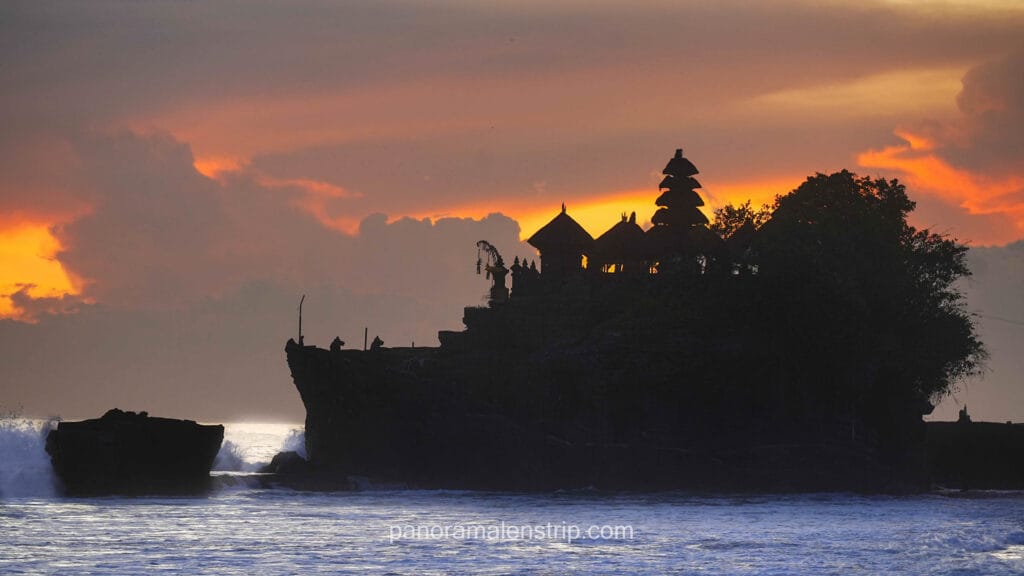 A dramatic silhouette of the Tanah Lot temple perched on a rocky ocean outcrop, set against a stunning, fiery orange and yellow sunset sky with dark clouds. Crashing ocean waves are visible at the base of the dark rock formation.