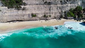 Pristine white sand beach nestled beneath a massive limestone cliff with turquoise ocean waves rolling ashore, featuring tiny silhouettes of tourists in Nusa Penida, Bali.