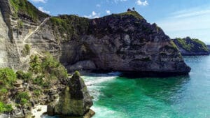 Dramatic white limestone cliffs with lush green tops meeting bright blue ocean waves in Nusa Penida, Bali, featuring a small wooden hut on the highest peak.