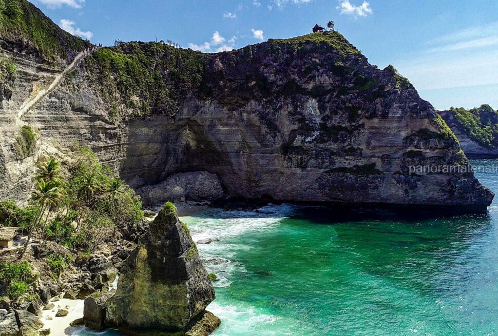 Dramatic white limestone cliffs with lush green tops meeting bright blue ocean waves in Nusa Penida, Bali, featuring a small wooden hut on the highest peak.