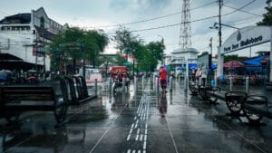 A wet pedestrian walkway on Malioboro Street in Yogyakarta after a rainstorm, featuring a traditional becak, street benches, and a sign for Pasar Sore Malioboro.