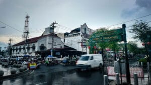 A bustling street corner in Yogyakarta showing a green directional sign for Polresta Yogyakarta, parked becaks, a moving white van, and historic white colonial-style buildings under a cloudy sky.