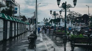 People wearing traditional Javanese clothing walking along a wet sidewalk lined with green kiosks and ornate street lamps in Yogyakarta, Indonesia.