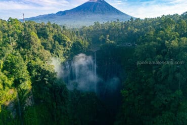 Aerial view of Tumpak Sewu waterfall cascading into a lush green jungle ravine, with the Mount Semeru volcano towering in the background in East Java, Indonesia.