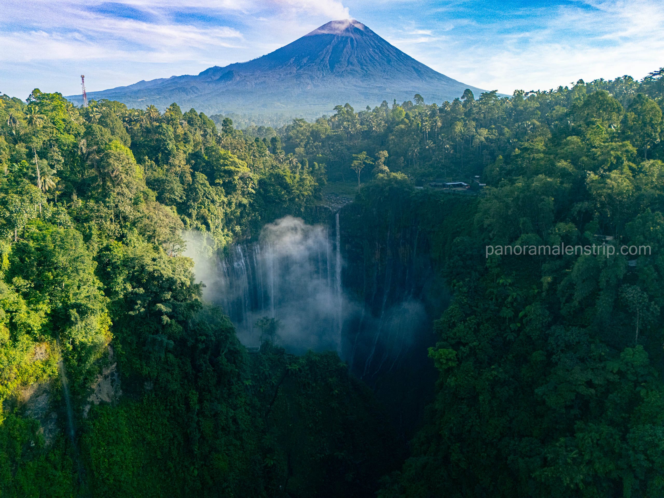 Aerial view of Tumpak Sewu waterfall cascading into a lush green jungle ravine, with the Mount Semeru volcano towering in the background in East Java, Indonesia.