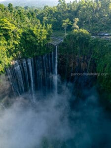 Aerial view of Tumpak Sewu Waterfall cascading down a lush, green, semi-circular cliff into a deep, misty gorge in East Java, Indonesia.