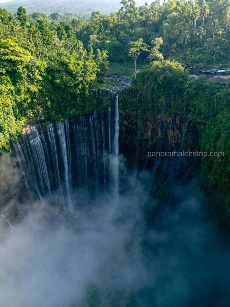 Aerial view of Tumpak Sewu Waterfall cascading down a lush, green, semi-circular cliff into a deep, misty gorge in East Java, Indonesia.