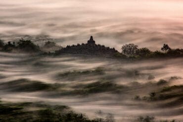Long exposure photography of Borobudur Temple emerging from a sea of thick morning mist at sunrise.