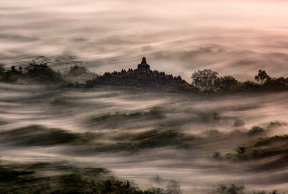 Long exposure photography of Borobudur Temple emerging from a sea of thick morning mist at sunrise.