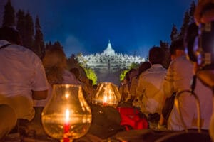 A wide night view of hundreds of pilgrims sitting in rows facing the illuminated Borobudur Temple for Vesak day.
