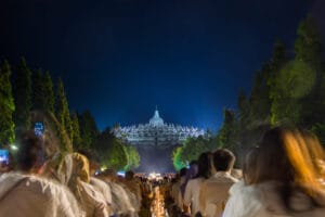 A wide night view of hundreds of pilgrims sitting in rows facing the illuminated Borobudur Temple for Vesak day.