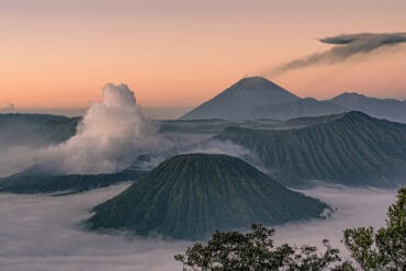 Active Mount Bromo emitting smoke next to Mount Batok and Mount Semeru, rising above a thick sea of morning clouds during sunrise in East Java, Indonesia.