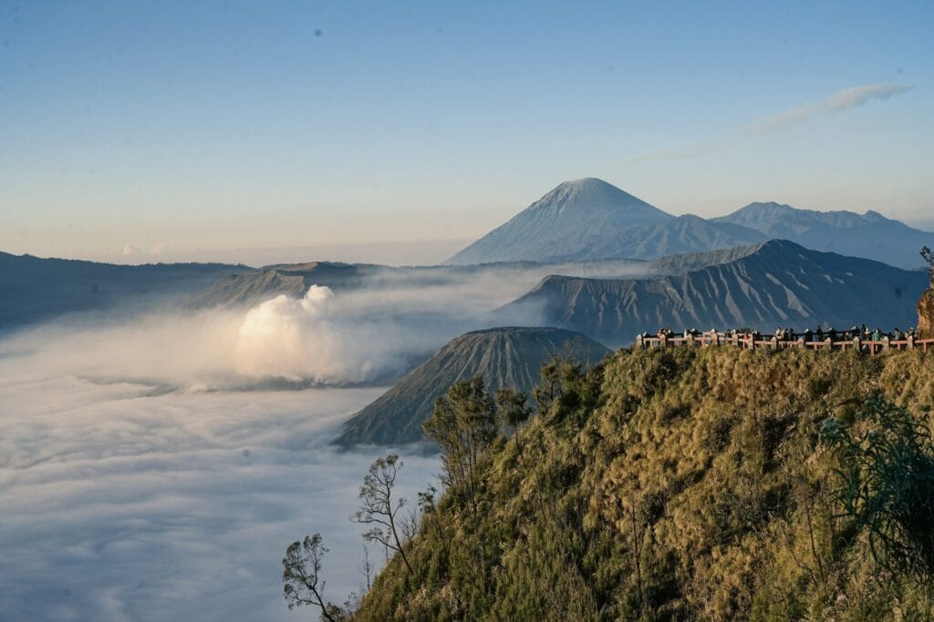 A crowd of tourists standing on a high-altitude viewing platform looking out over Mount Bromo, Mount Batok, and a sea of fog under a clear blue morning sky in Indonesia.