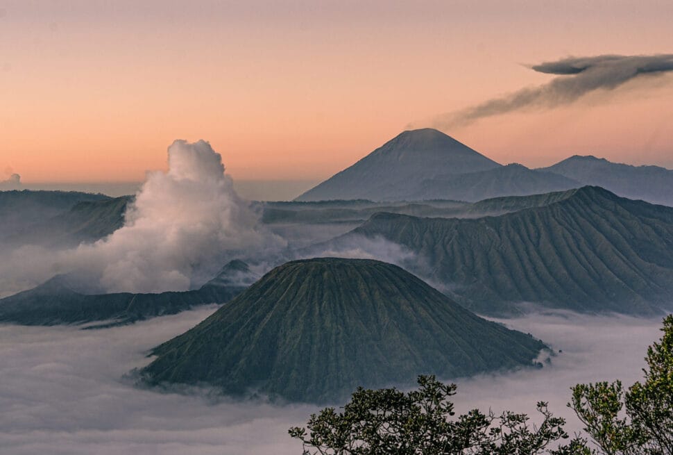Active Mount Bromo emitting smoke next to Mount Batok and Mount Semeru, rising above a thick sea of morning clouds during sunrise in East Java, Indonesia.
