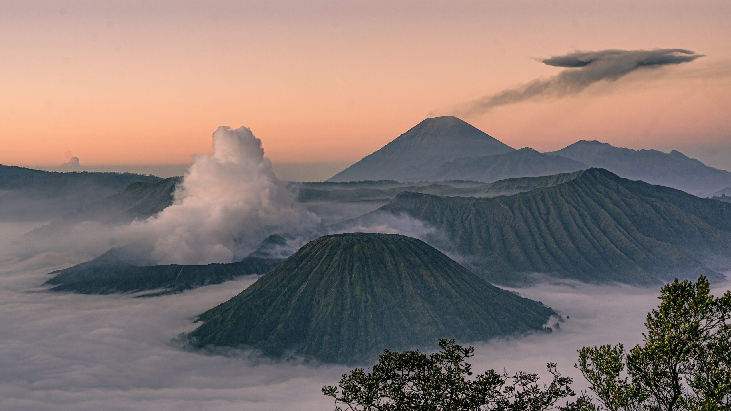 Active Mount Bromo emitting smoke next to Mount Batok and Mount Semeru, rising above a thick sea of morning clouds during sunrise in East Java, Indonesia.