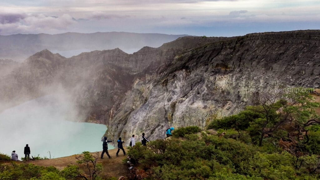 Tourists hiking along a dirt path on the rim of the Kawah Ijen crater in East Java, showcasing steep, textured volcanic rock cliffs on the right and the smoky turquoise acidic lake on the left.