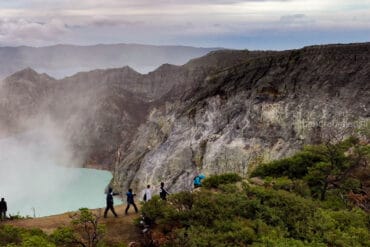Tourists hiking along a dirt path on the rim of the Kawah Ijen crater in East Java, showcasing steep, textured volcanic rock cliffs on the right and the smoky turquoise acidic lake on the left.