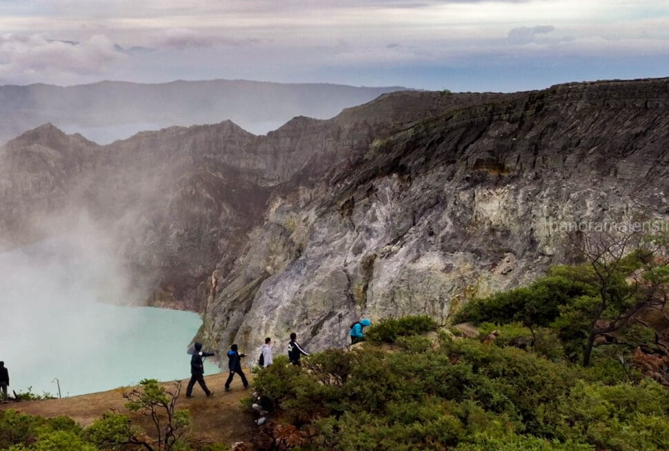 Tourists hiking along a dirt path on the rim of the Kawah Ijen crater in East Java, showcasing steep, textured volcanic rock cliffs on the right and the smoky turquoise acidic lake on the left.