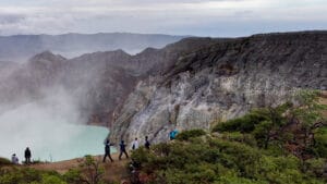 Tourists hiking along a dirt path on the rim of the Kawah Ijen crater in East Java, showcasing steep, textured volcanic rock cliffs on the right and the smoky turquoise acidic lake on the left.