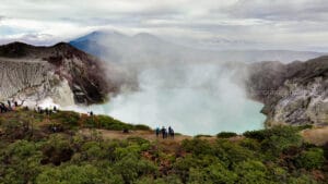 Wide panoramic view of the turquoise acidic crater lake at Mount Ijen in East Java, Indonesia, surrounded by steep rocky cliffs and thick white sulfur smoke, with tourists observing from the rim.