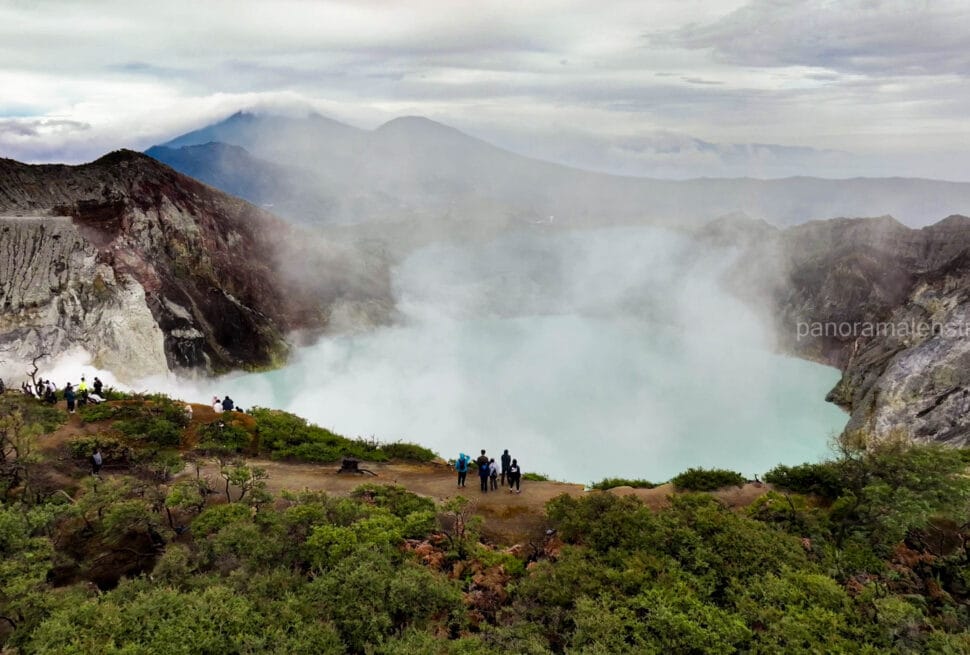 Wide panoramic view of the turquoise acidic crater lake at Mount Ijen in East Java, Indonesia, surrounded by steep rocky cliffs and thick white sulfur smoke, with tourists observing from the rim.